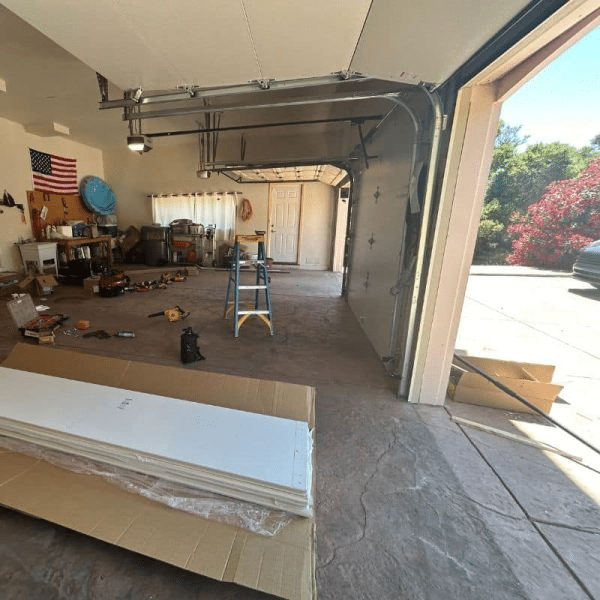 Inside a cluttered garage undergoing work: new white garage door panels stacked on cardboard boxes, a yellow stepladder, tools scattered on the floor, and the existing door angled halfway open.